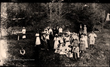 A group of casually posed people in a wooded area, two holding Concord College banners. Other information includes "HDN Coll. early 20th Century, S. Trail 2002". 