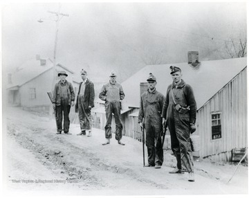 Five miners with guns and buckets stop on the road in front of houses for a picture. 