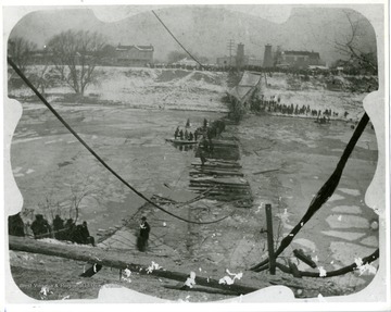 Cables and wooden bridge has fallen into the Elk River. People line the shore and bank of river to watch the rescuing of people from the bridge.  