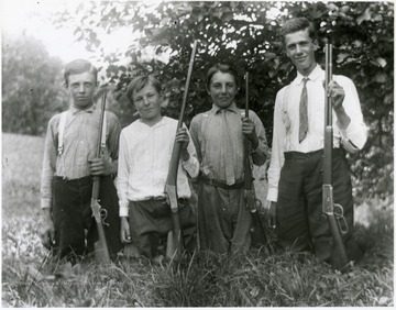 Group portrait of four boys kneeling in the grass, each holding a gun.