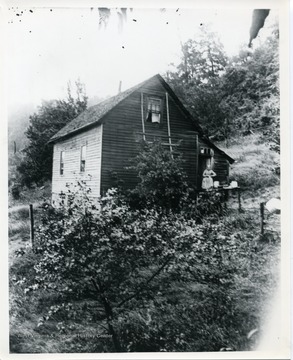 An unidentified woman standing on a porch in Helvetia, West Virginia. The wooden two-story house is surrounded by trees.