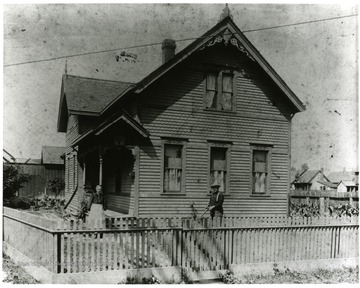 An elderly woman is standing in her front yard while an older gentleman is cutting the grass. Their two-story wooden house is on a small plot surrounded by a wooden picket fence.  Location of home or identification of individuals is unknown.