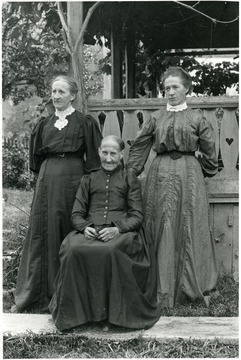 The three ladies are in dresses posing for a portrait outside.