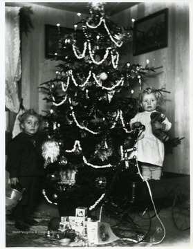 Two cousins who are standing near their Christmas tree with their presents.