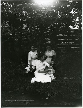 Three women sitting in forested area looking at postcard foldout book.  Helvetia, W. Va.