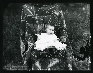 A young baby sitting in a chair which is covered with a quilt.