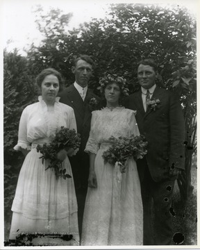 Two ladies and two gentlemen are standing together near a tree. The two ladies are holding flowers.  Possibly a wedding portrait.