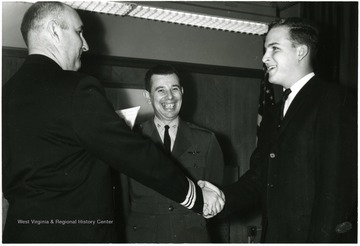 A young man shakes hands with what appears to be a man from the Military, another man from the Air Force stands in the background. 