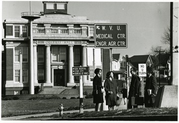 Students chat in front of Oglebay Hall.  Signs point to the Medical Center, the Engineering and Agriculture Center, General Hospital, Star City and the Bureau of Mines.