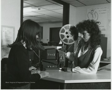 Right: Tina M. Parton, Audiovisual Technical Assistant and  Left: Catherine J. Groves, Library Technical Assistant in  Colson Hall.  Trina explains the operation of a 16 mm projector to Catherine.