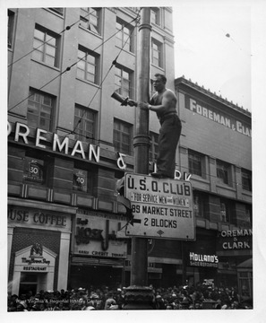 A man stands on a sign up a pole with a fire siren in his hand.